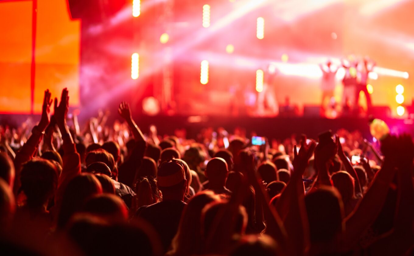 Audience with raised hands watching a live concert under colorful stage lighting.